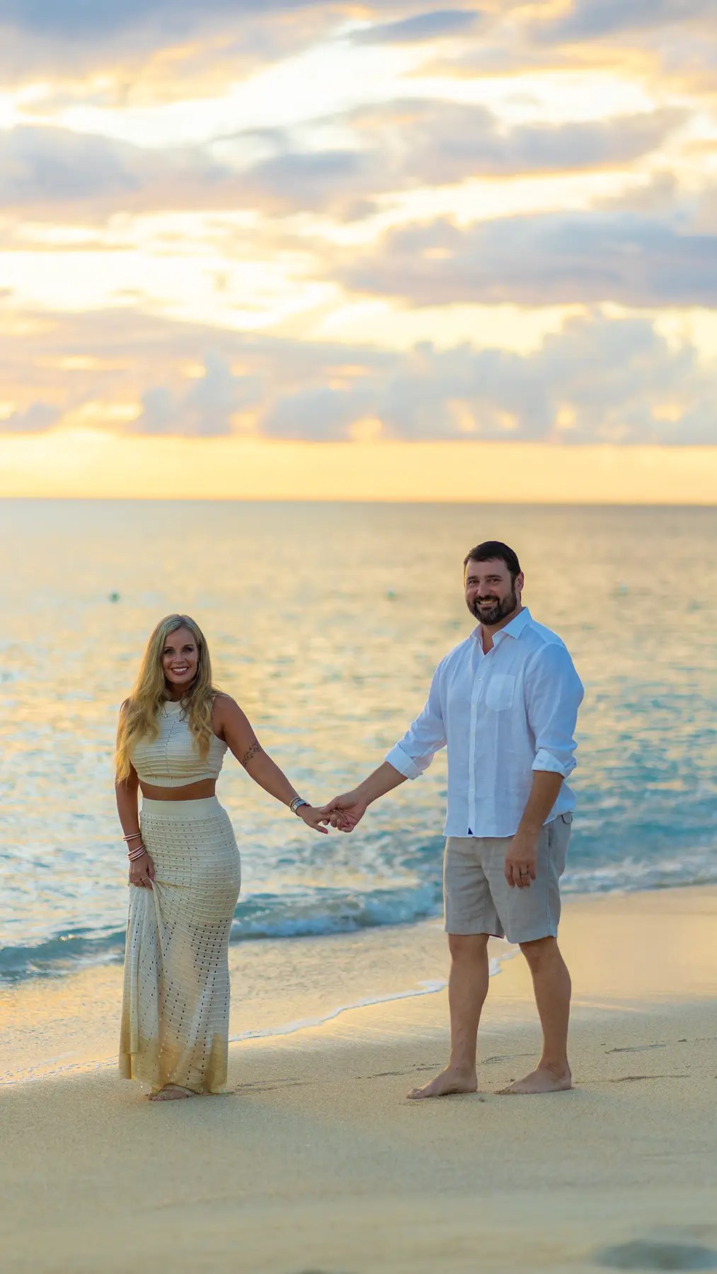 Couple holding hands on a Caribbean beach at sunset, dressed in light summer clothing, with ocean waves and vibrant clouds in the background