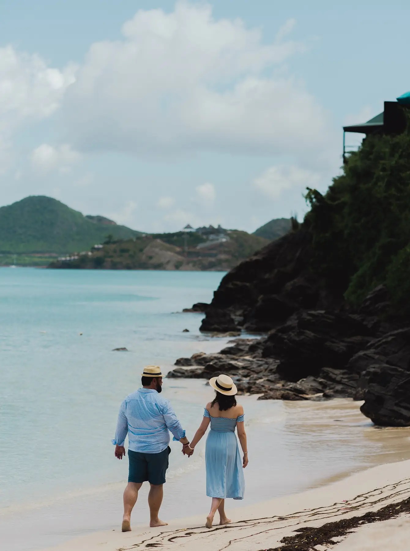 A couple holding hands while walking along a sandy beach, with mountains in the background and a clear blue sky in Antigua.