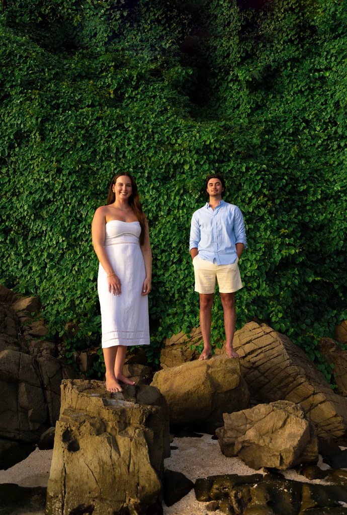 A woman in a white dress and a man in a light blue shirt stand on rocks by the beach, surrounded by lush green foliage.