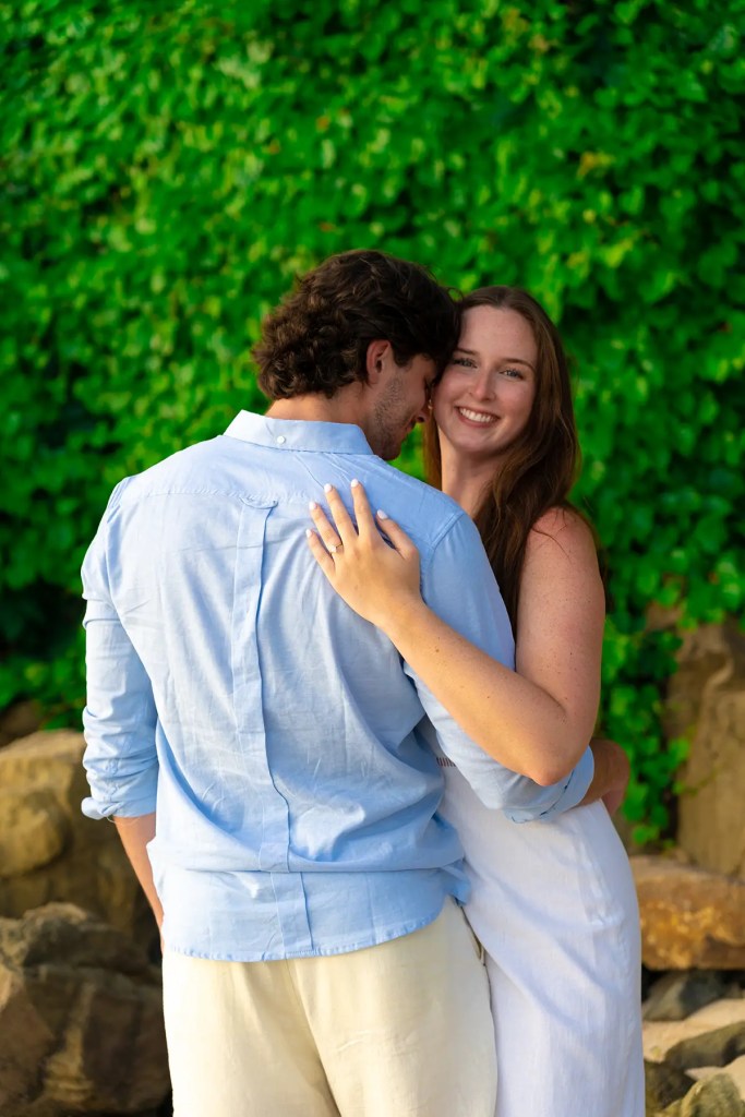 A couple embracing outdoors, with a lush green backdrop. The woman is smiling at the camera while the man leans in close, creating an intimate moment.