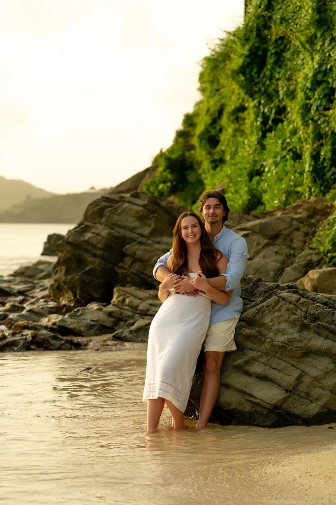 A couple standing together on a beach, with the woman wearing a white dress and the man in a light blue shirt. They are smiling and embracing near a rocky area by the water.